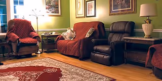 A cozy living room with green walls, wooden flooring, and a large patterned rug. The room features a red upholstered sofa with pillows, a matching armchair, a dark brown recliner, two wooden side tables with lamps, and framed artwork on the walls.