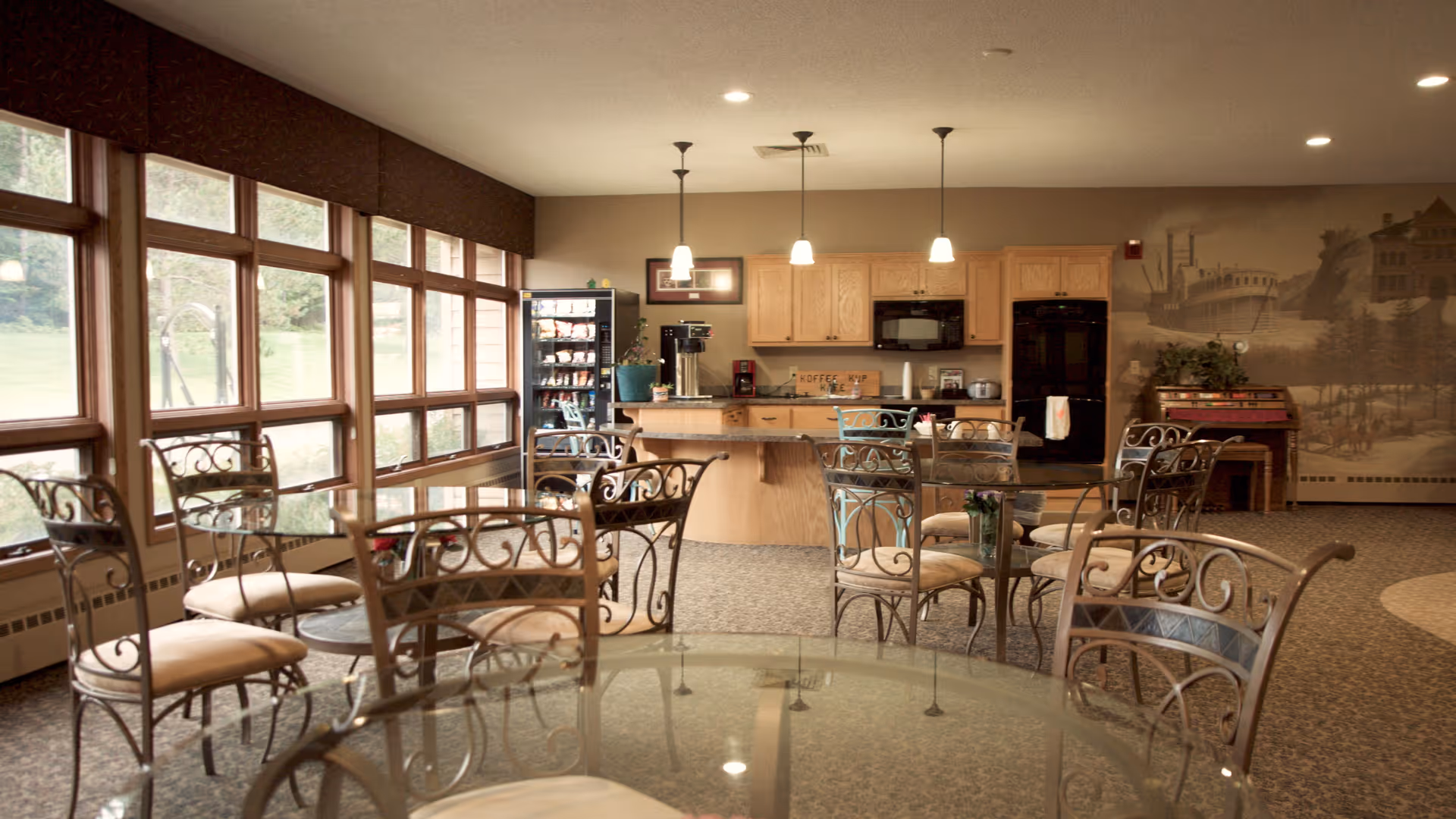 Sunlit communal dining area with glass-top tables and wrought-iron chairs facing a kitchenette and large windows.