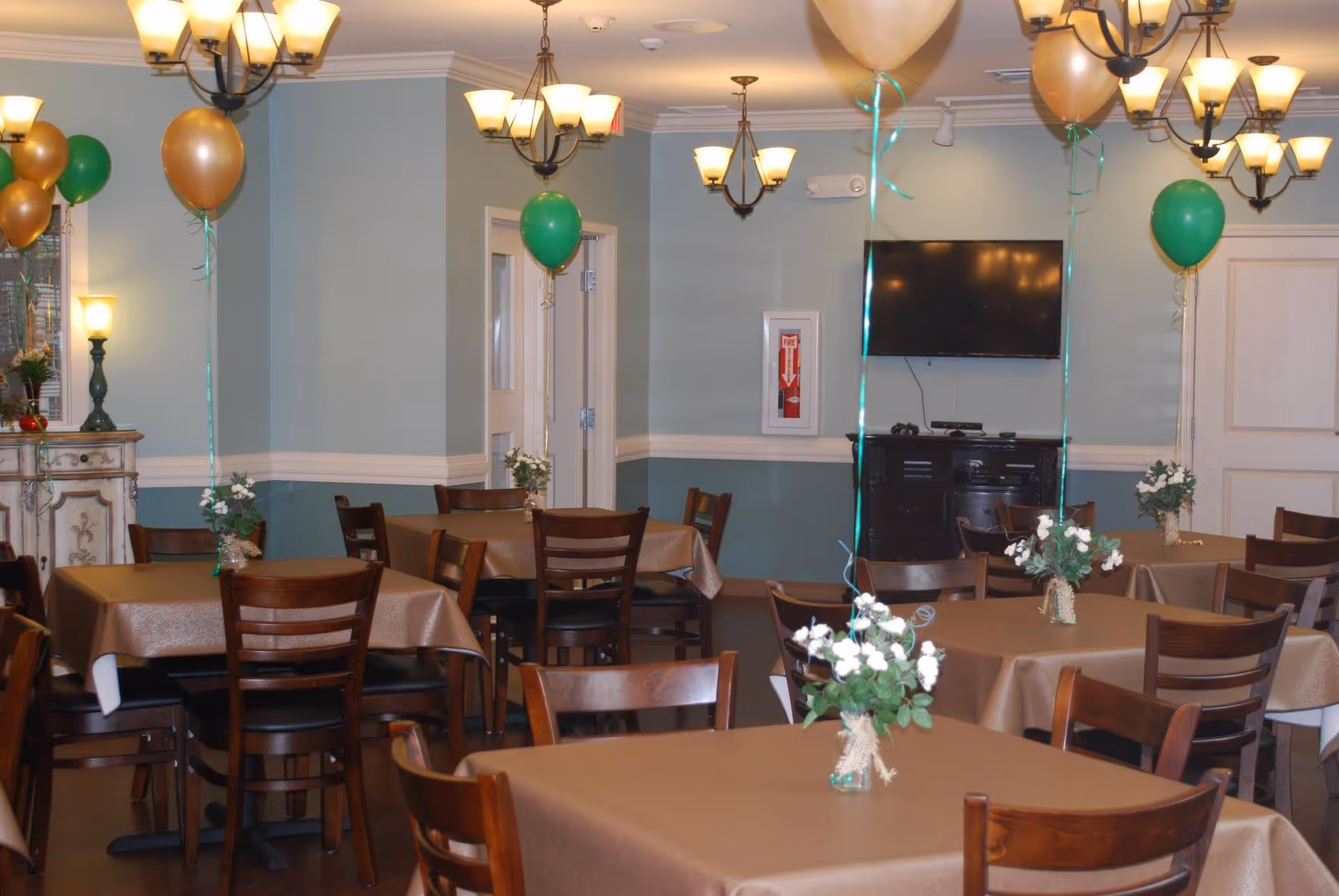 A dining room with multiple tables covered in brown tablecloths, each decorated with small flower arrangements and balloons in gold and green colors. The room has soft blue walls, several ceiling light fixtures, a wall-mounted TV, and a cabinet beneath it.