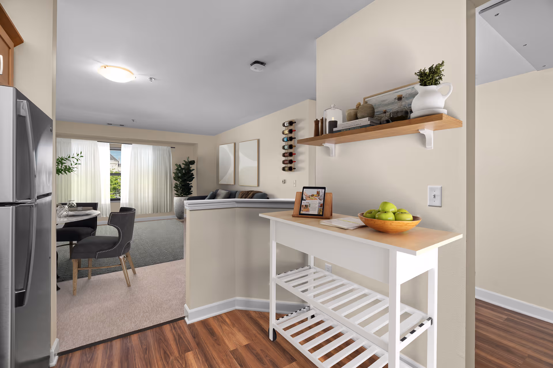 Interior view of a senior living facility showing a kitchen area with a stainless steel refrigerator on the left, a white wooden console table with a bowl of green apples and decorative items on the right, and an open living and dining area in the background with a round dining table, chairs, a sofa, wall art, and large windows with sheer curtains letting in natural light.