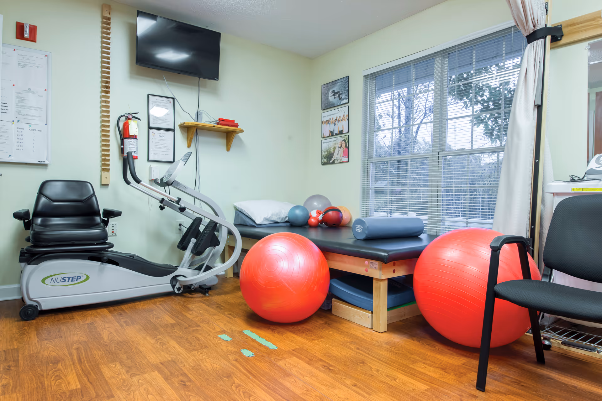 A small exercise room with a recumbent exercise bike, two large red exercise balls, a padded therapy table with various smaller exercise balls and a foam roller on it, a black chair, and a large window with blinds letting in natural light.