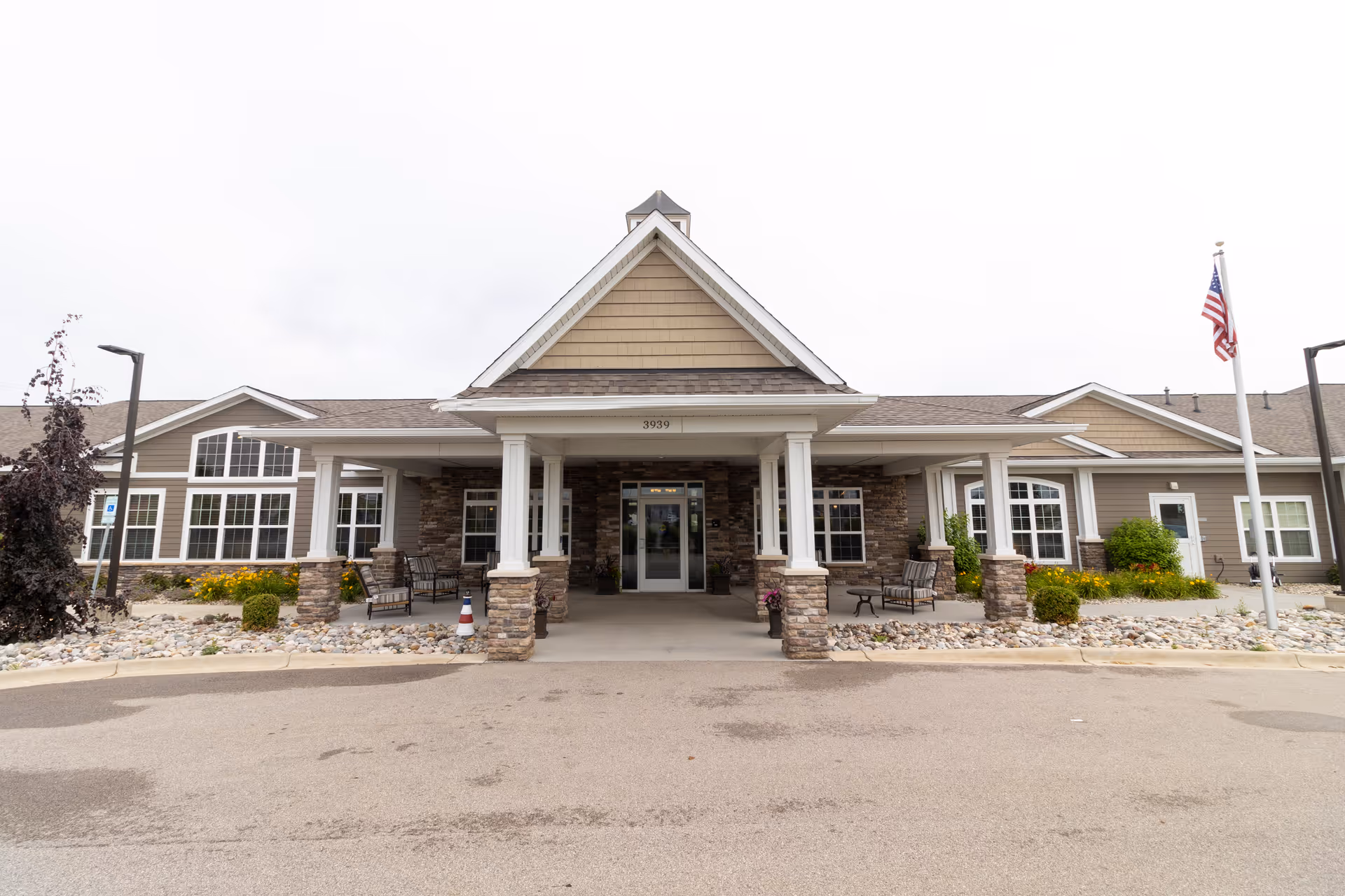 Front entrance of a single-story assisted living building with a covered portico, stone columns, benches and an American flag.
