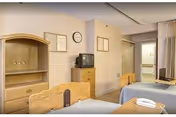 Interior view of a senior living facility bedroom with two beds, wooden furniture including a dresser and a shelving unit, a small television on top of the dresser, a wall clock, and framed pictures on the wall. A doorway leads to a hallway with handrails.