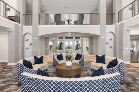 Bright two-story communal lounge with curved patterned sofas and navy pillows arranged around a central round coffee table beneath an upper balcony.