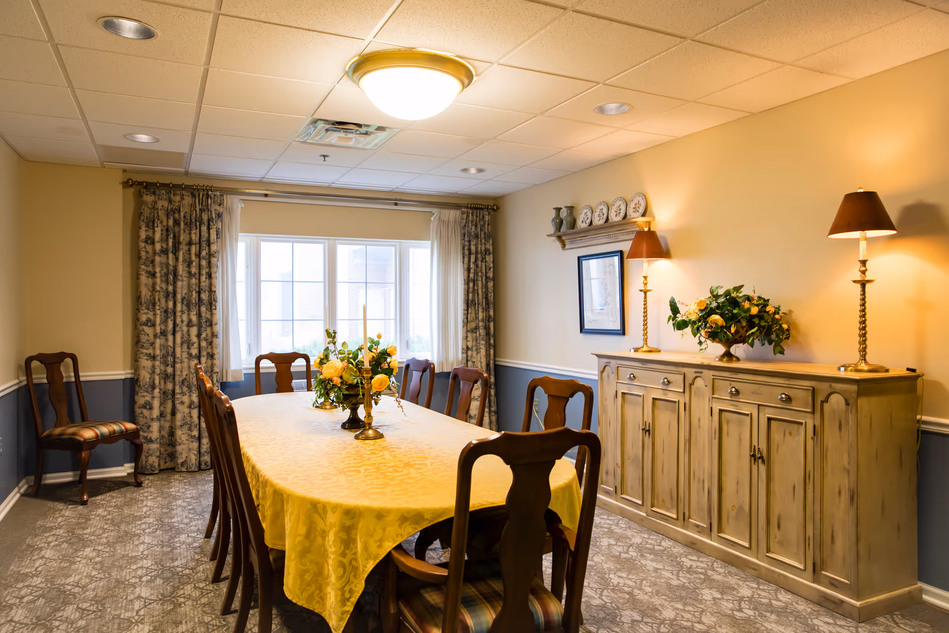 A warmly lit dining room with a long table covered by a yellow tablecloth, surrounded by wooden chairs with patterned cushions. The table is decorated with a floral centerpiece and candlesticks. A large window with floral curtains lets in natural light. On the right side, there is a wooden sideboard with two lamps and a flower arrangement, and a shelf above it holding decorative plates.