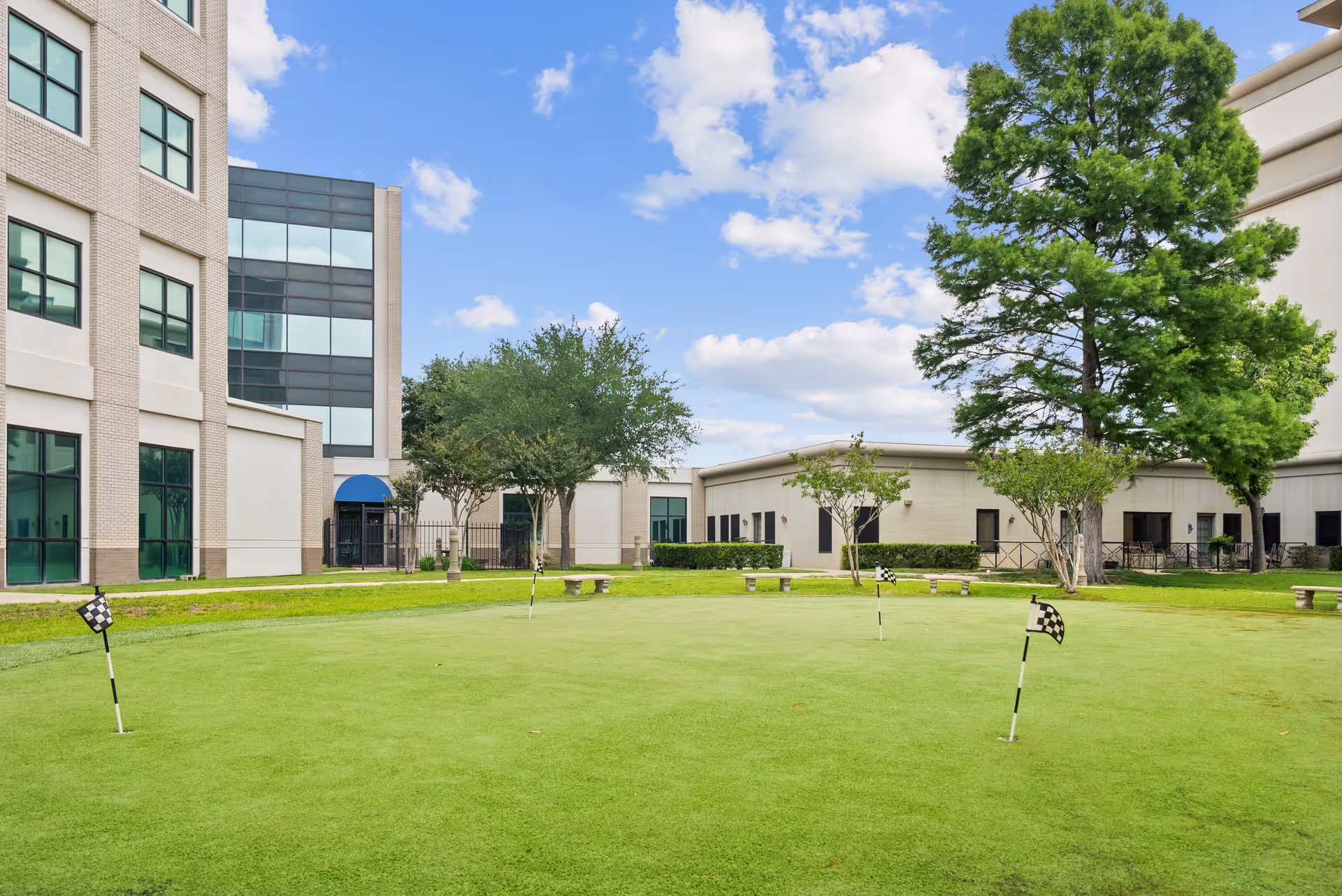 Outdoor putting green with several small checkered flags on poles, surrounded by benches, trees, and buildings under a partly cloudy blue sky.