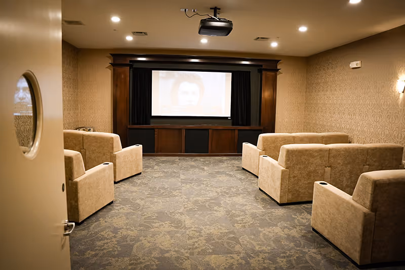 A small movie theater room with beige upholstered recliner chairs arranged in two rows facing a large screen. The room has patterned carpet, wallpapered walls, and a ceiling-mounted projector.