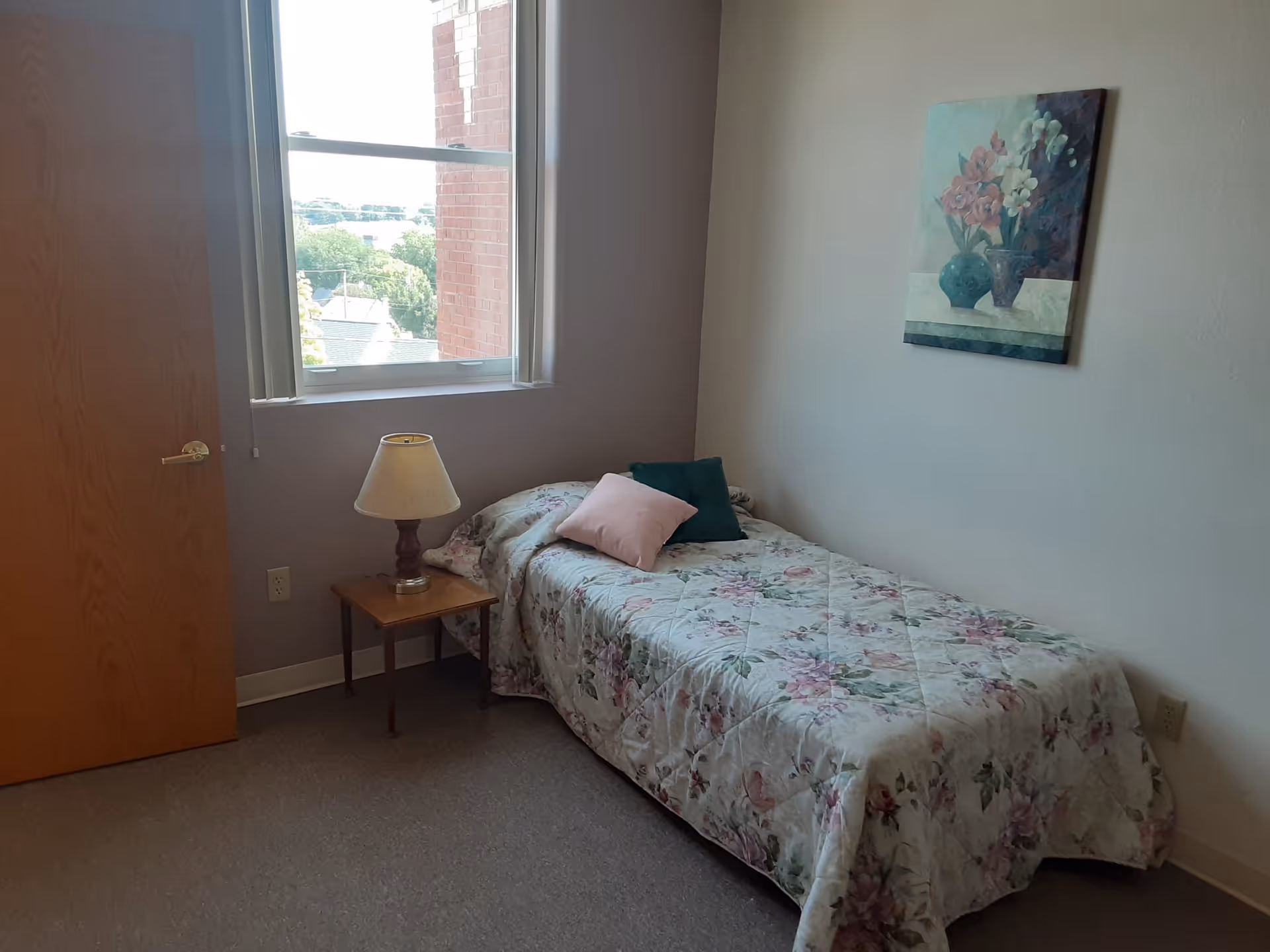 A small bedroom with a single bed covered in a floral bedspread and two pillows, one pink and one green. Next to the bed is a wooden nightstand with a lamp. There is a window with a view of a brick building and greenery outside. A floral painting hangs on the wall above the bed.