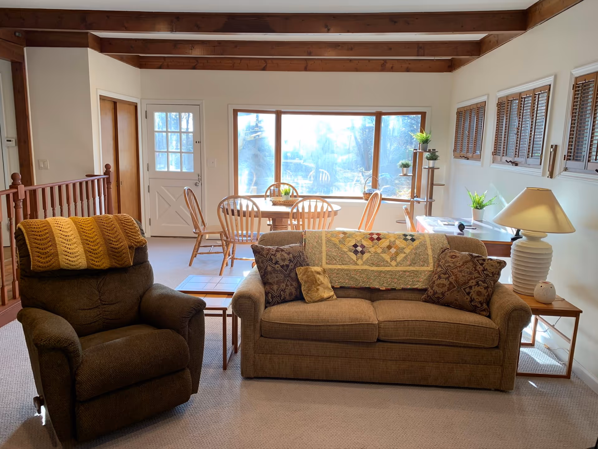 A cozy living room area with a brown armchair draped with a yellow knitted blanket and a beige sofa adorned with patterned cushions and a quilt. Behind the seating area is a wooden dining table with chairs near a large window letting in natural light. The room features wooden ceiling beams, a white door, several small windows with wooden blinds, and a side table with a lamp and potted plants.