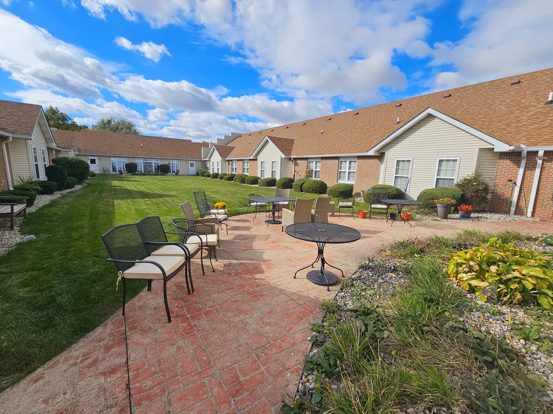 Sunny courtyard with metal tables and chairs on a brick patio surrounded by single-story residential buildings and landscaping.