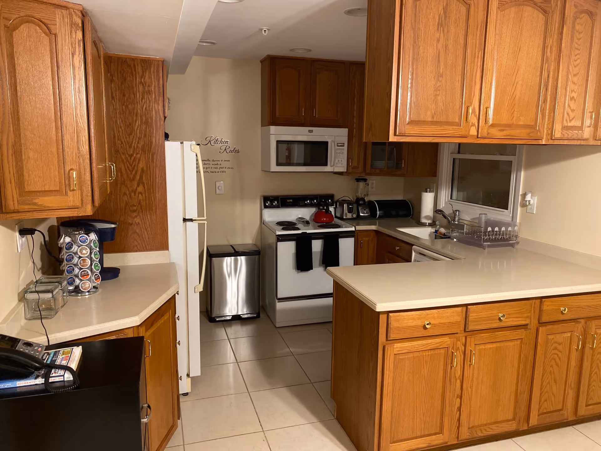 A kitchen with wooden cabinets, white countertops, a white refrigerator, a white stove with a red kettle on it, a microwave above the stove, a stainless steel trash can, a coffee maker with coffee pods, a blender, a paper towel holder, and a dish drying rack next to a window.