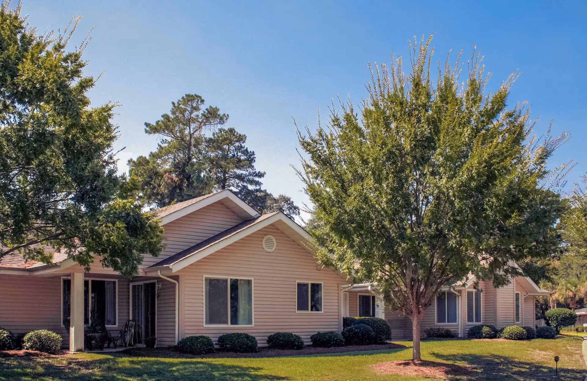 Exterior view of a single-story residential building with beige siding, surrounded by green trees and shrubs under a clear blue sky.