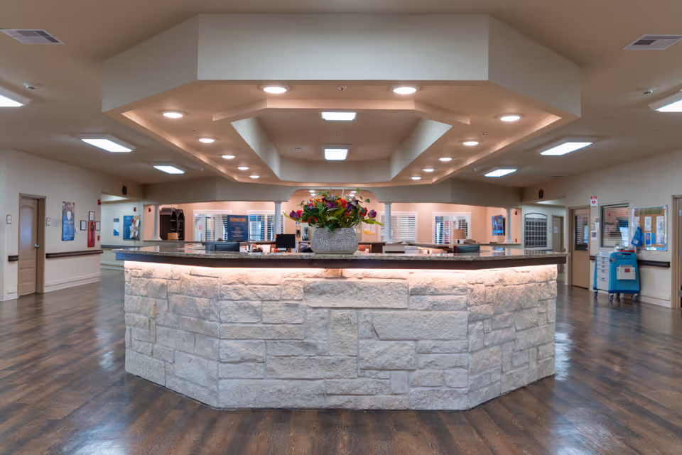 Interior view of a senior living facility reception area with a large stone-faced front desk topped with a granite countertop and a vase of flowers. The ceiling features recessed lighting and a unique architectural design. The room has wood flooring and several doors and windows along the walls.