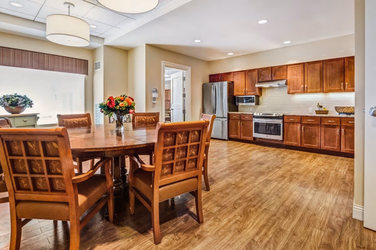 A spacious kitchen and dining area with wooden flooring and cabinetry. The kitchen features stainless steel appliances including a refrigerator, oven, and microwave. A round wooden dining table with six cushioned chairs is centered in the room, with a vase of colorful flowers on the table. Large windows with blinds allow natural light to fill the space.