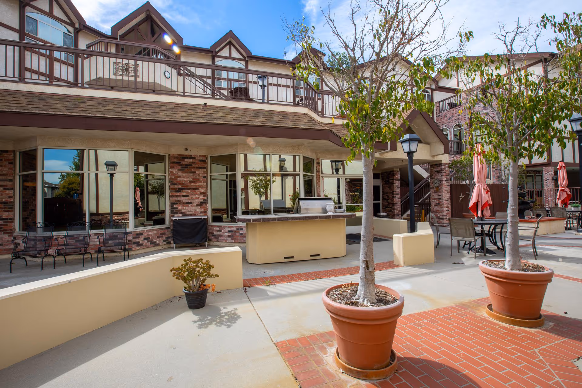 Outdoor courtyard area of Lexington Assisted Living featuring potted trees, patio tables with umbrellas, benches, and a built-in grill. The building has brick and beige walls with large windows and a second-floor balcony.