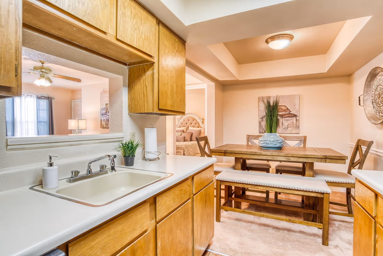 View of a kitchen sink with wooden cabinets and a white countertop, looking into a dining area with a wooden table, cushioned bench, and chairs. The dining area has a decorative plant centerpiece, wall art, and a ceiling light fixture. A bedroom is partially visible through an open doorway.