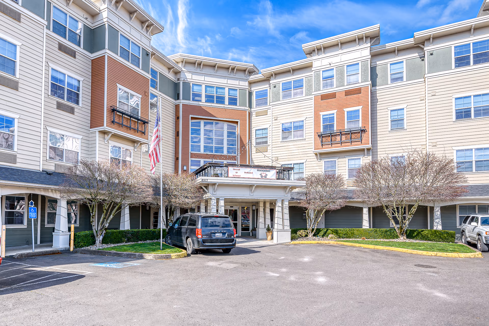 Front exterior of a multi-story senior living building with an entrance canopy, parked cars, and an American flag.