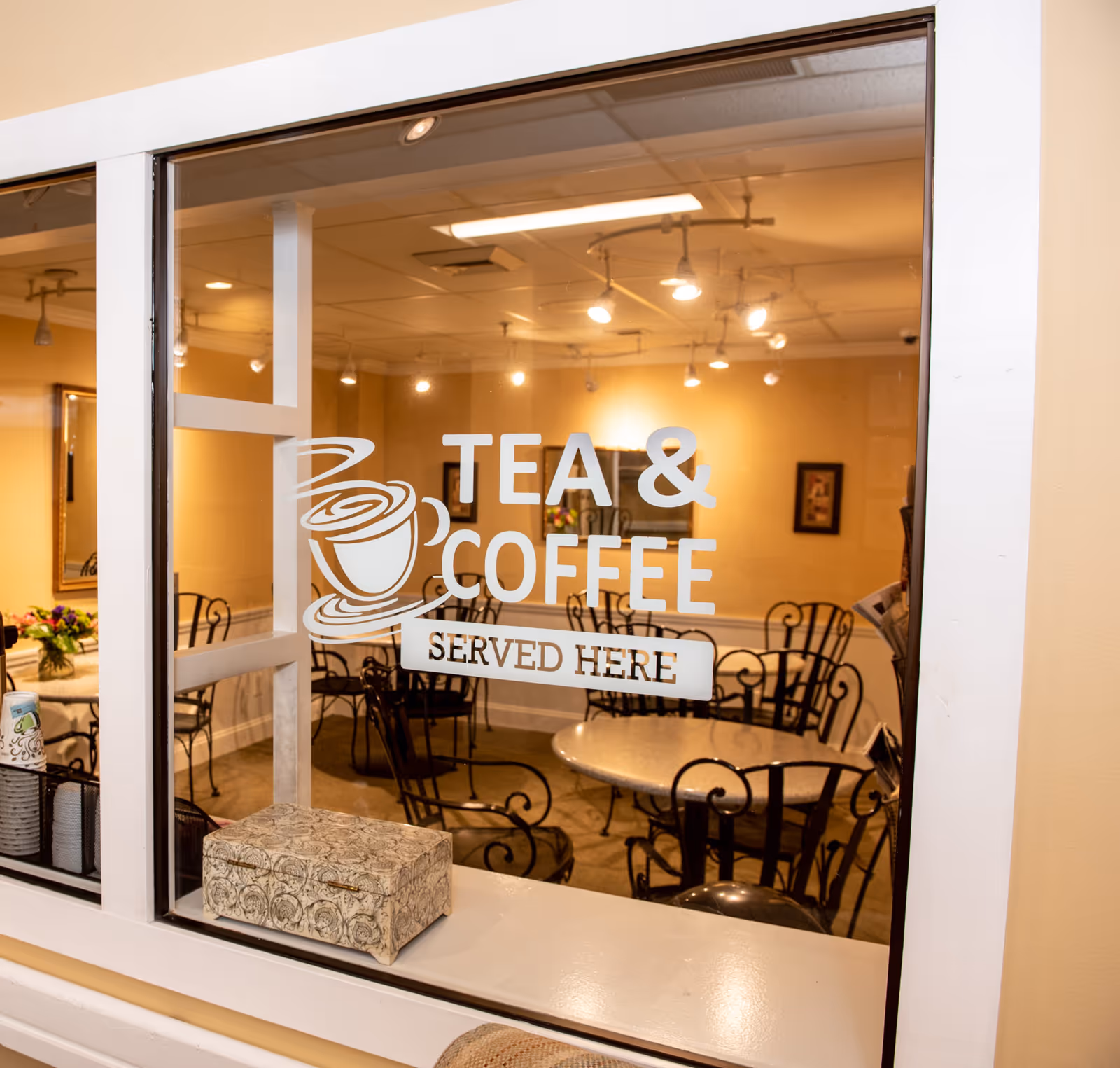 View through a glass window into a cozy room with several round tables and black wrought iron chairs. The glass has a decal with a steaming cup icon and the words 'TEA & COFFEE SERVED HERE'. The room has warm lighting and framed pictures on the walls.