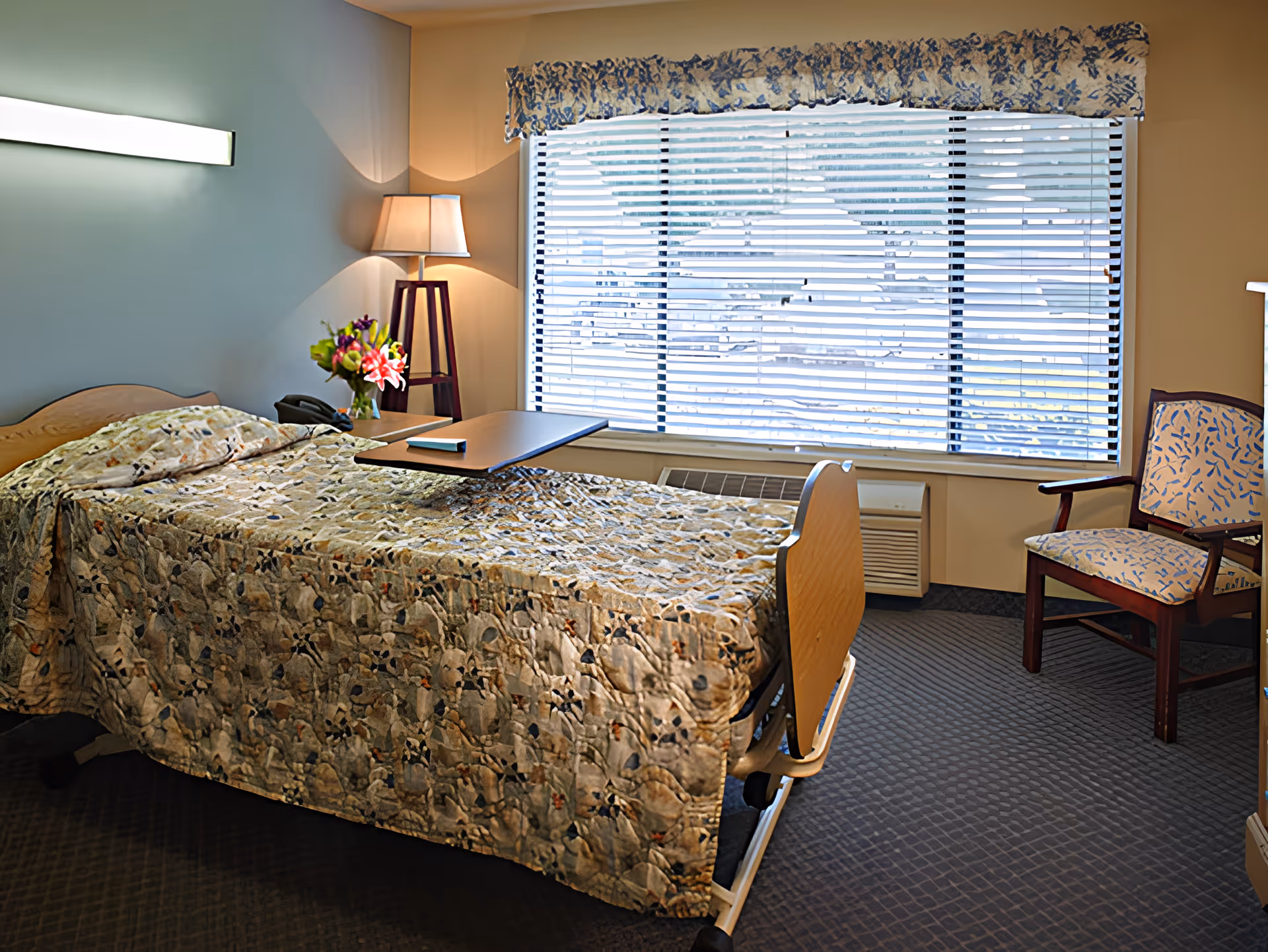 A single hospital-style bed with a patterned quilt in a well-lit room. Next to the bed is a small table with a telephone and a vase of flowers. A floor lamp stands in the corner near a large window with blinds and a decorative valance. There is a cushioned chair with wooden arms and legs near the window, and an air conditioning unit below the window.