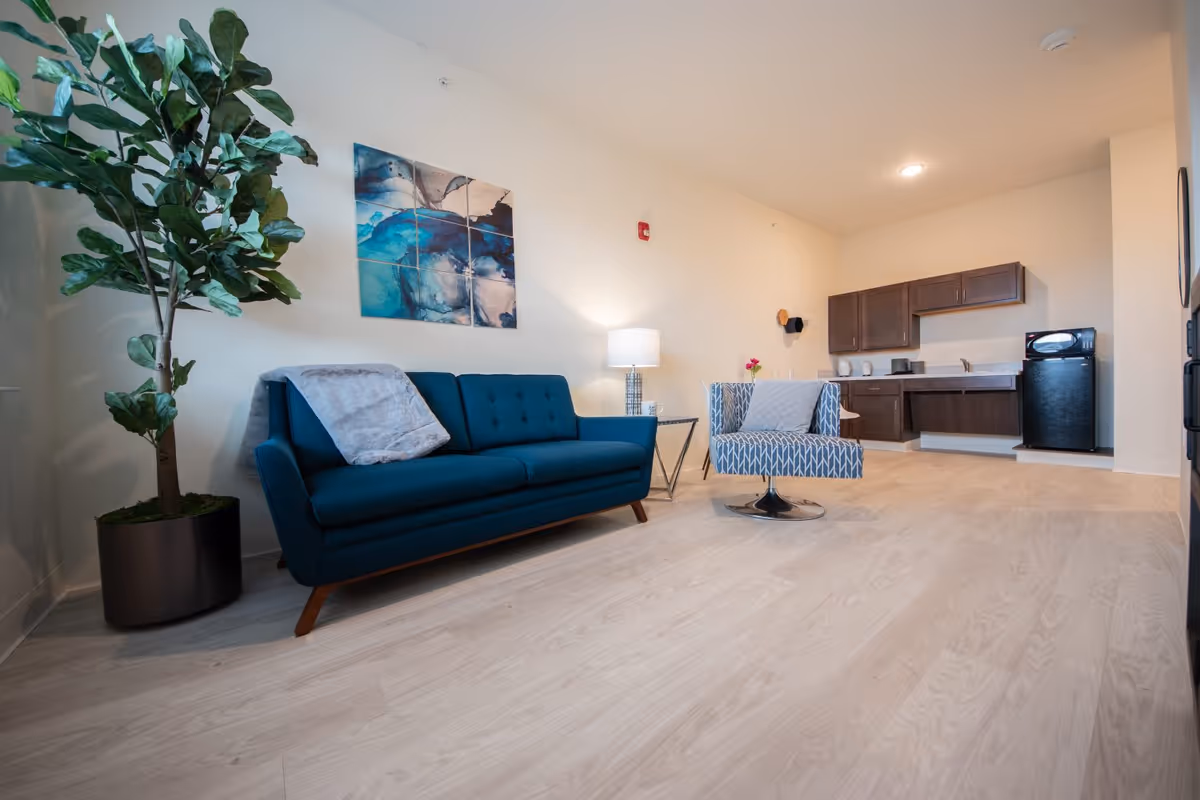A modern living area in a senior living facility featuring a blue sofa with a gray throw blanket, a patterned armchair with a gray pillow, a side table with a lamp, a large potted plant, and a kitchenette with dark wood cabinets, a black mini fridge, and a microwave.