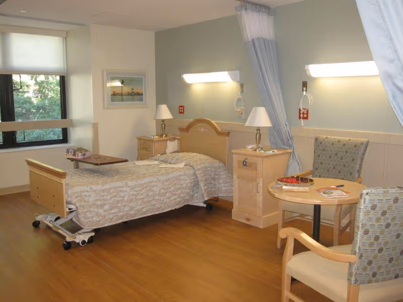 A well-lit patient room in a healthcare facility featuring a single hospital bed with a beige patterned bedspread, two bedside tables with lamps, a small round table with two chairs, a window with a view of greenery, and light blue privacy curtains partially drawn.