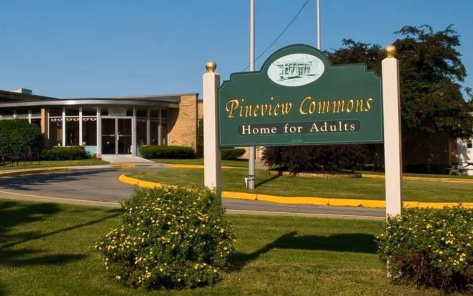Green entrance sign reading 'Pineview Commons Home for Adults' in front of a single-story facility building with driveway and landscaping.
