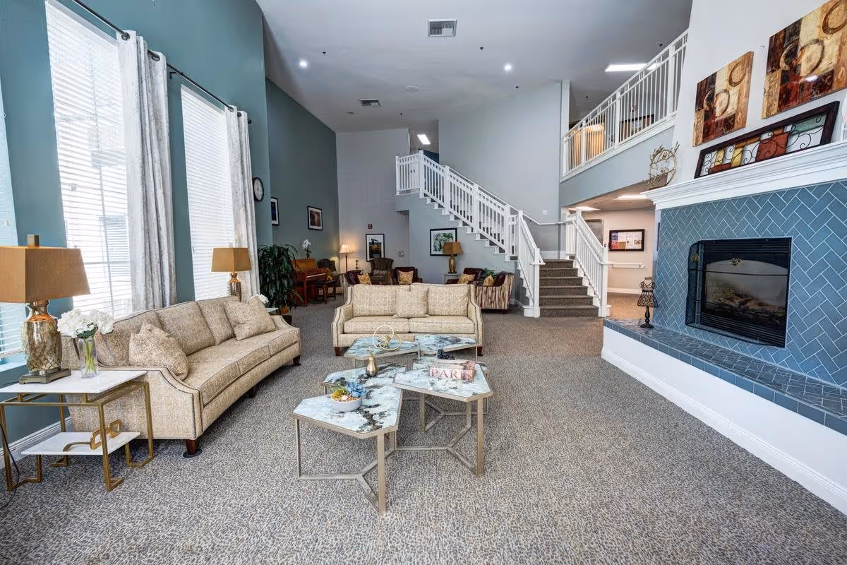 A spacious and well-lit living room area in Creston Village featuring two beige sofas, a set of three marble-top coffee tables, a blue-tiled fireplace, and a staircase leading to an upper floor. The room has large windows with white curtains, beige carpet flooring, and decorative lamps and artwork on the walls.