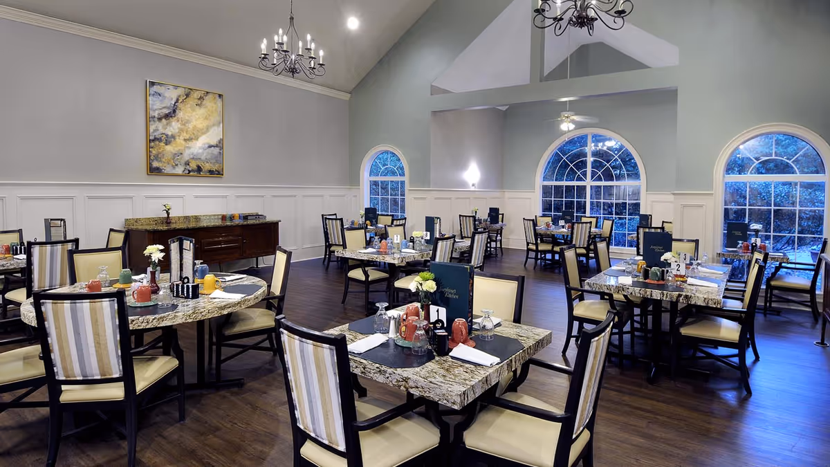 A spacious dining room with multiple tables set for meals, featuring granite tabletops and chairs with striped upholstery. The room has large arched windows, high ceilings with chandeliers, and a sideboard against the wall with a decorative painting above it.