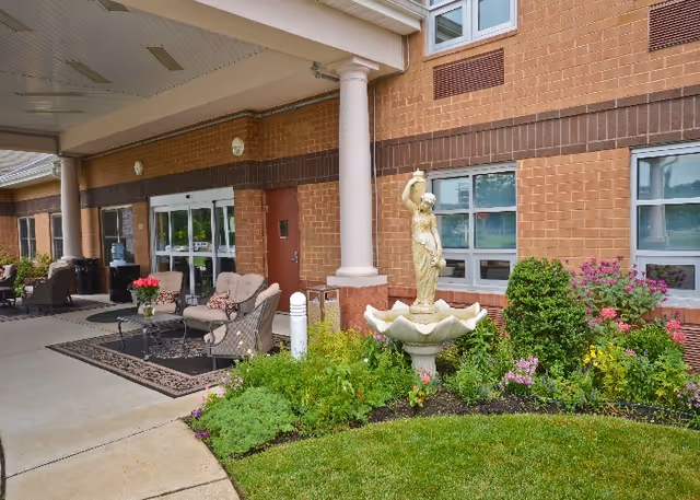 Outdoor seating area with cushioned chairs and a table on a rug near the entrance of a brick building. There is a decorative statue fountain surrounded by green plants and flowers in front of the building.