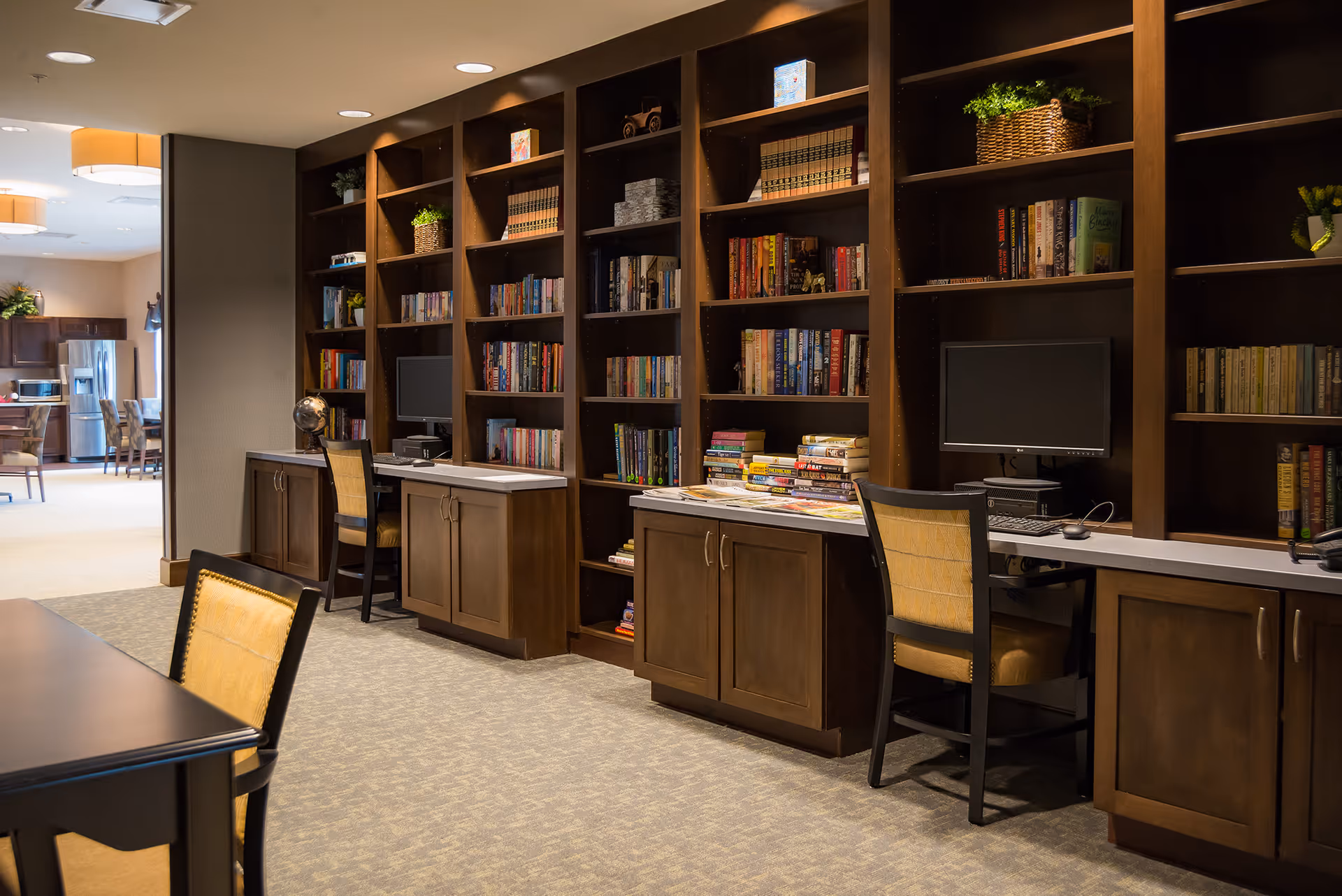 Interior view of a senior living facility library and computer area with wooden bookshelves filled with books, two computer workstations with chairs, and a table with chairs in the foreground. The background shows a dining area with tables and chairs and a kitchen with stainless steel appliances.