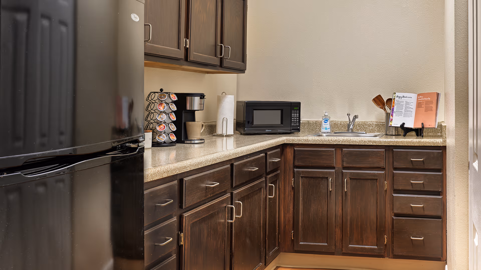 A kitchen area with dark wooden cabinets and drawers, a black refrigerator on the left, a coffee maker with a pod holder, a microwave, a paper towel holder, a sink with a faucet, hand soap, and an open cookbook on a stand on the countertop.