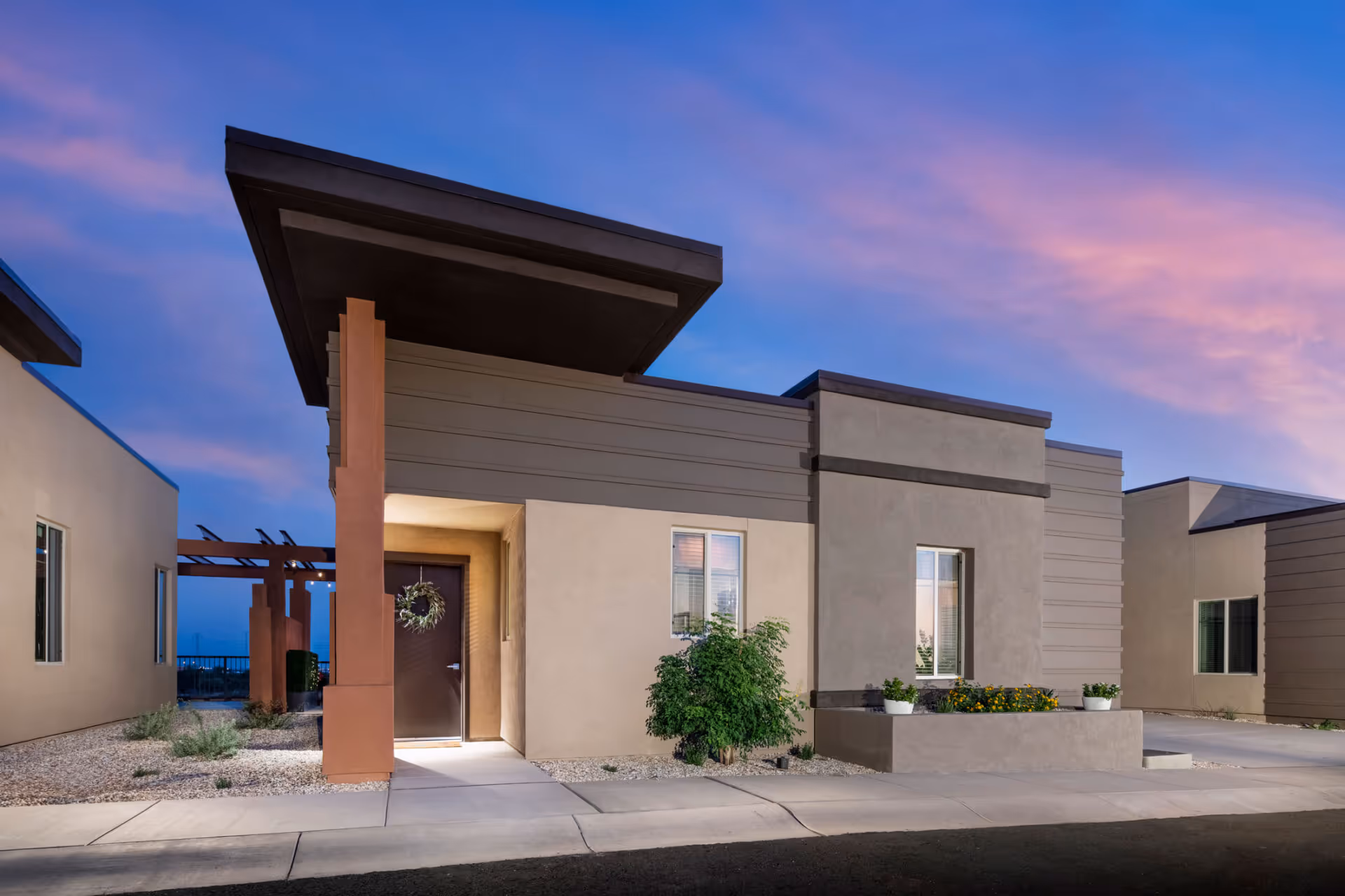 Exterior view of a modern single-story building at sunset with a covered entrance, beige and brown walls, a door decorated with a wreath, windows, and some landscaping including bushes and potted plants.