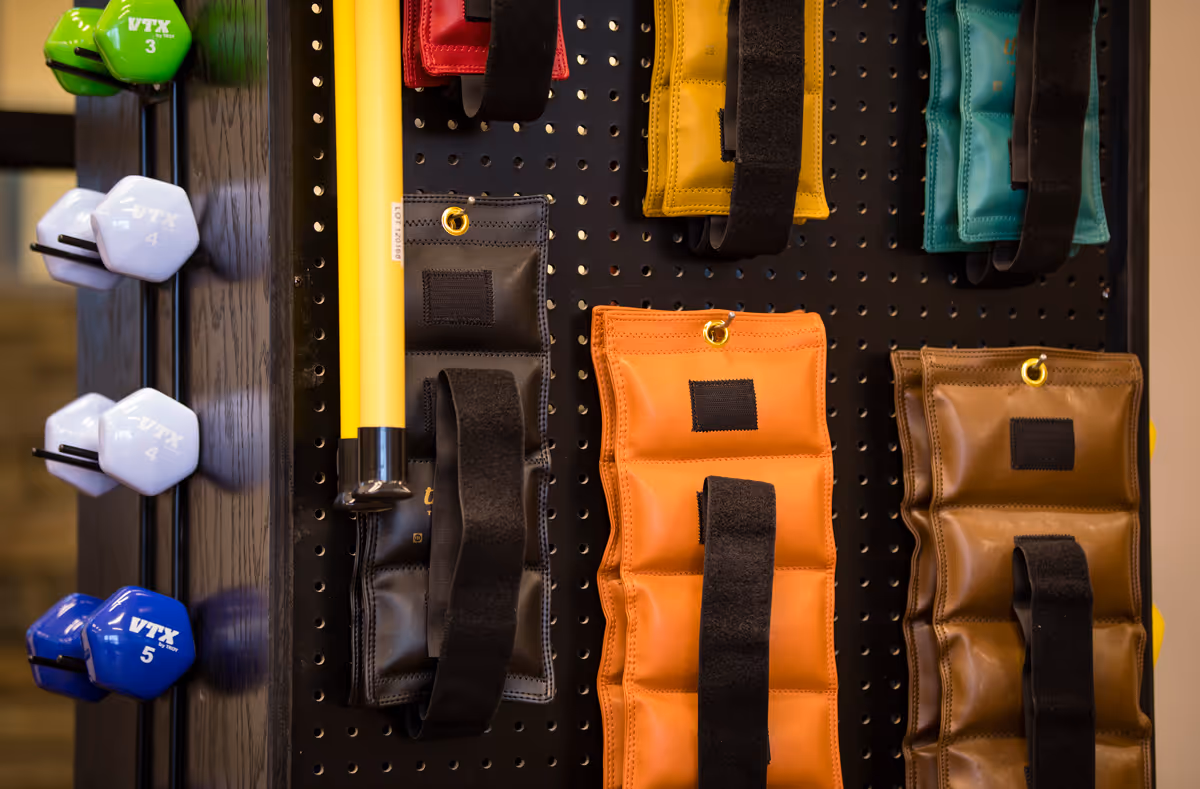A close-up view of a fitness equipment rack with colorful weighted wrist or ankle straps hanging on a pegboard and various dumbbells of different weights arranged on the side.