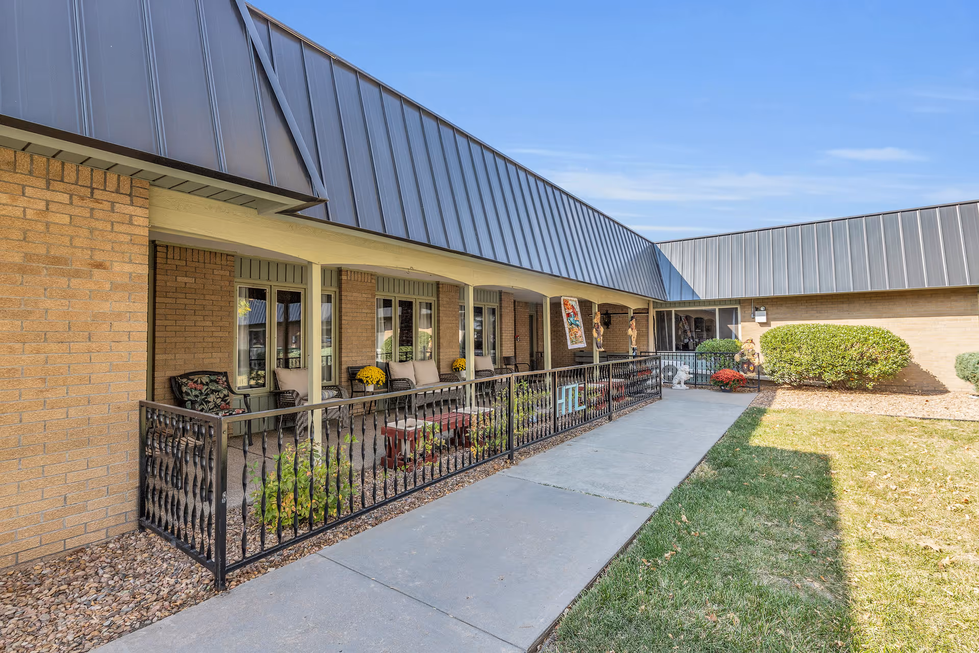 Outdoor patio area of a senior living facility with a covered walkway, several chairs, small tables, potted plants, and a black metal railing. The building has light brown brick walls and a metal roof. There is a concrete pathway and a grassy area adjacent to the patio.