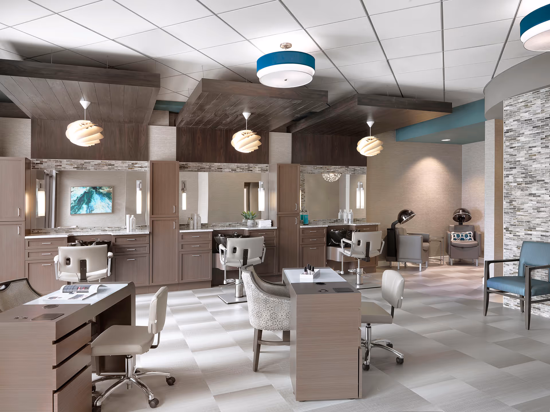 A modern salon area in a senior living facility featuring multiple styling stations with mirrors, chairs, and cabinetry. The space includes a manicure table with chairs in the foreground and hair drying chairs with hooded dryers in the background. The room has a light color palette with wood accents, pendant lighting, and a tiled floor.