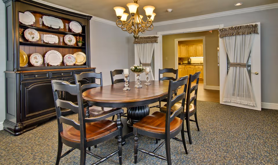 A dining room with a round wooden table surrounded by six wooden chairs with dark frames and brown seats. On the table are three decorative candle holders. Behind the table is a black wooden hutch displaying decorative plates. The room has carpeted flooring and a chandelier hanging from the ceiling. In the background, there is an open doorway leading to a kitchen with wooden cabinets and a coffee maker visible. The doorway has white curtains tied back with brown sashes.