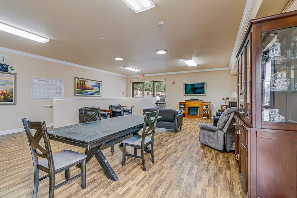 A spacious living room area in an assisted living facility featuring a long black table with four chairs, several armchairs and sofas arranged around a wooden electric fireplace with a flat-screen TV mounted above it. The room has wood flooring, beige walls with framed paintings, and a glass cabinet displaying decorative items on the right side. Large windows and a door at the back provide natural light and a view of an outdoor patio area.