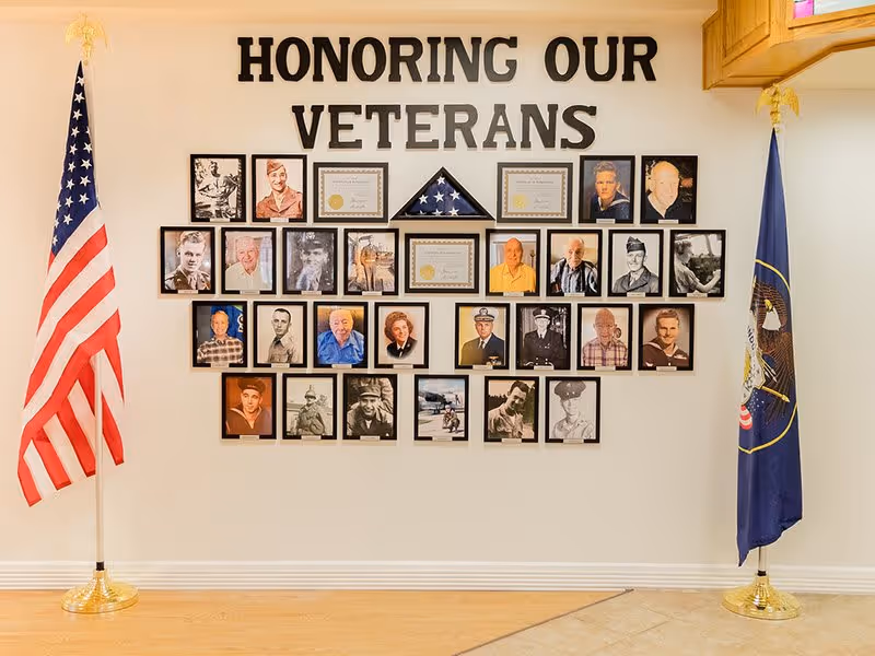 A wall display honoring veterans with framed photographs of veterans and certificates arranged around a folded American flag in a triangular case. The words 'HONORING OUR VETERANS' are displayed above the photos. An American flag stands on a pole to the left and a Utah state flag stands on a pole to the right.