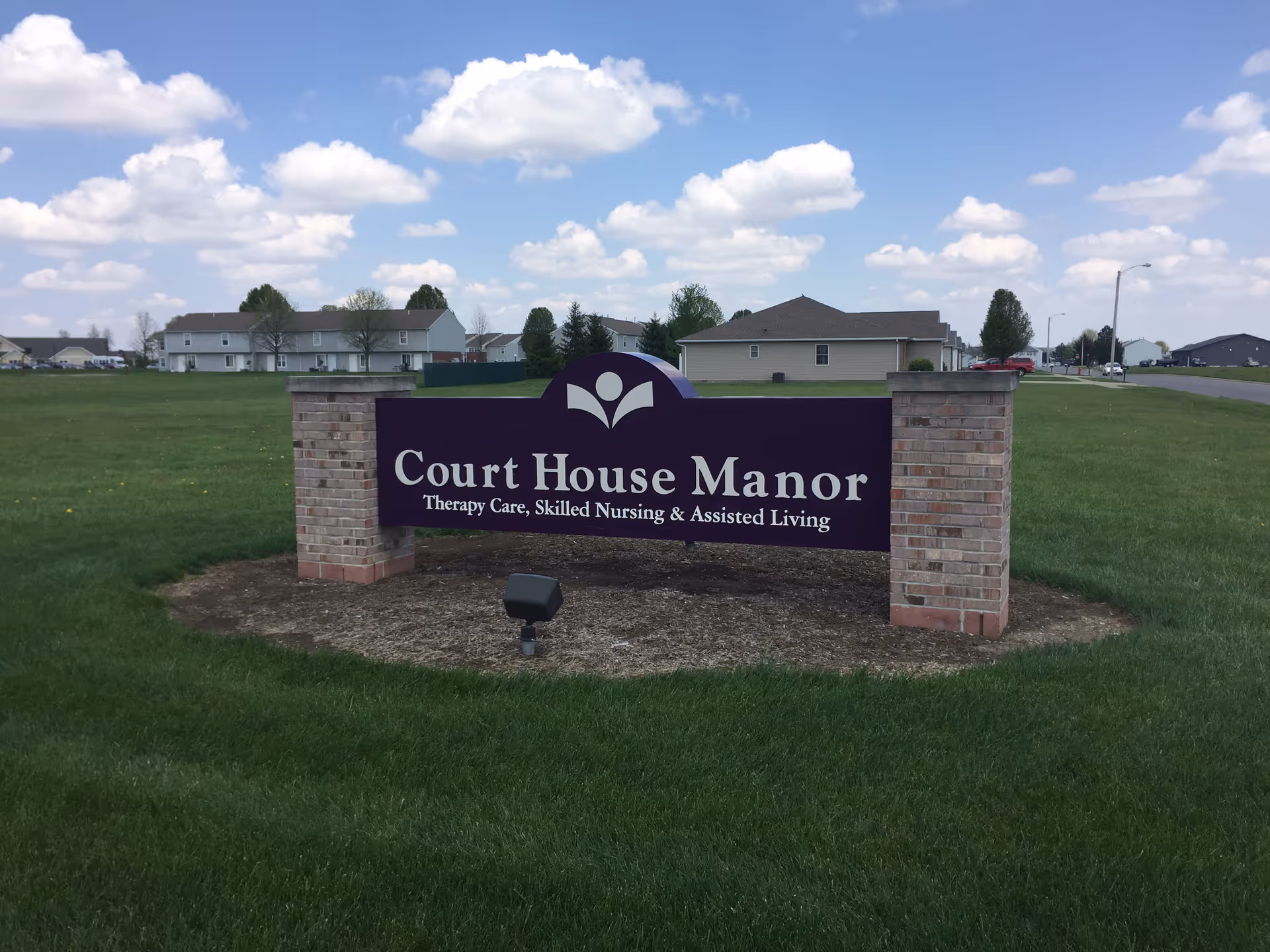 Outdoor view of a large purple sign with white text that reads 'Court House Manor Therapy Care, Skilled Nursing & Assisted Living' mounted between two brick pillars on a grassy lawn with houses and a partly cloudy sky in the background.