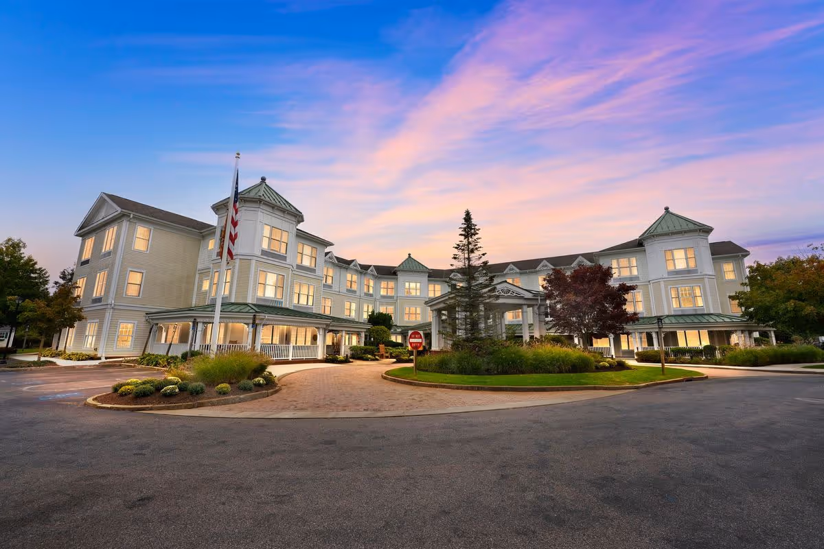 Large three-story senior living facility building with a circular drive, landscaped island, and flagpole at dusk.