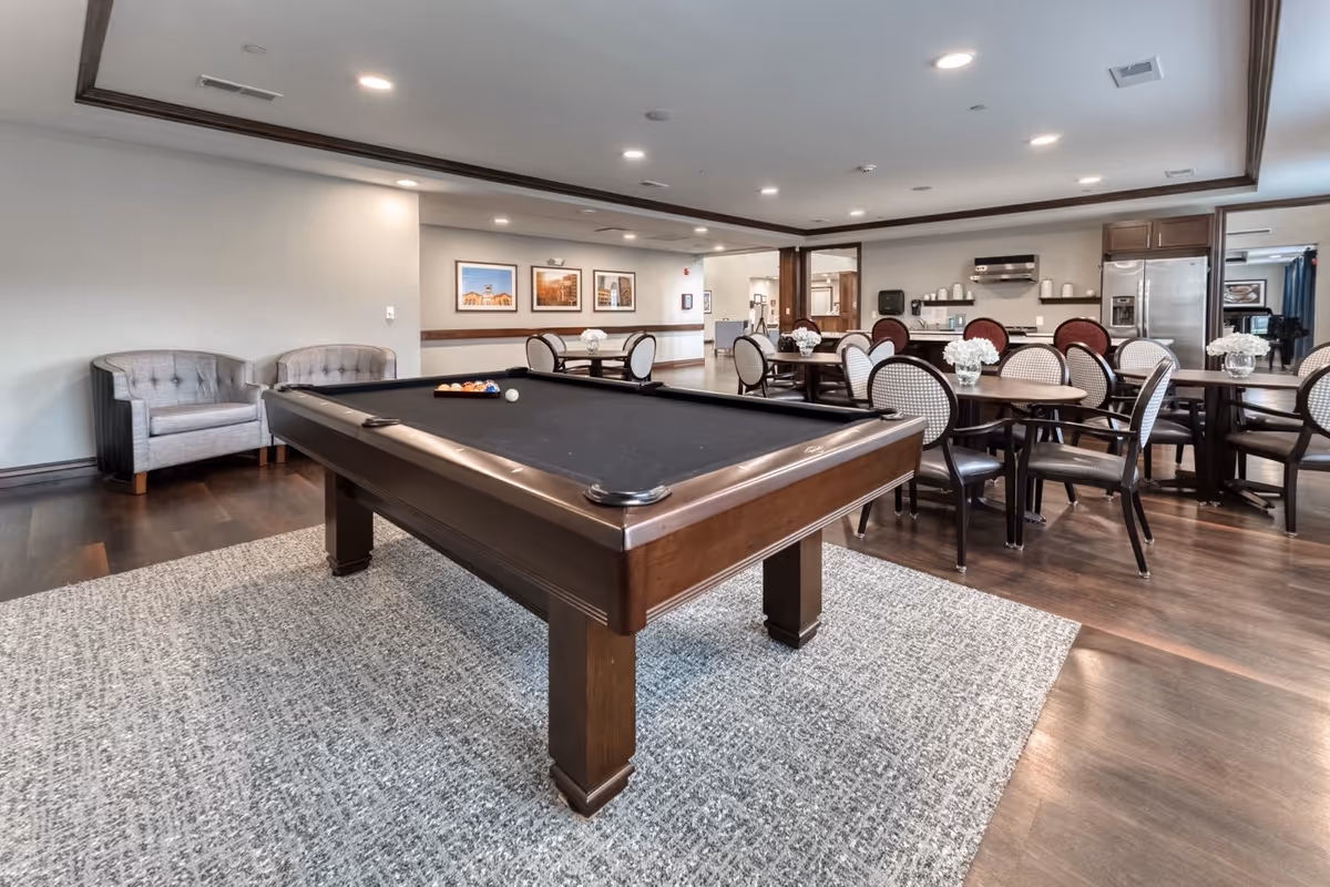 A senior living facility common area featuring a pool table with balls arranged for a game, several round tables with chairs, and a kitchenette area with a refrigerator and shelves. The room has hardwood floors, a large area rug under the pool table, and framed pictures on the wall.