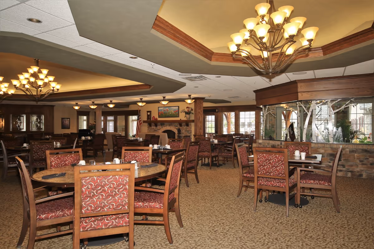 A spacious dining room with multiple round and square wooden tables surrounded by chairs with red patterned upholstery. The room features warm lighting from chandeliers and ceiling lights, a carpeted floor, and a stone fireplace with a painting and decorative items on the mantel. Large windows and glass panels with decorative tree designs allow natural light to enter the room.