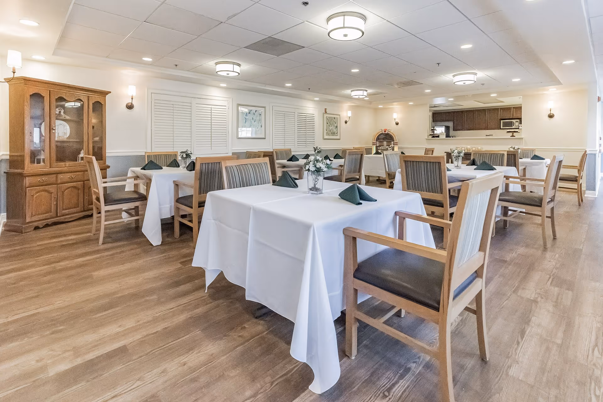 A bright dining room with several tables covered in white tablecloths, each set with folded green napkins and small floral centerpieces. Wooden chairs with cushioned seats surround the tables. The room features wood flooring, a wooden cabinet, wall sconces, framed artwork, and ceiling lights. A kitchen area is visible in the background.