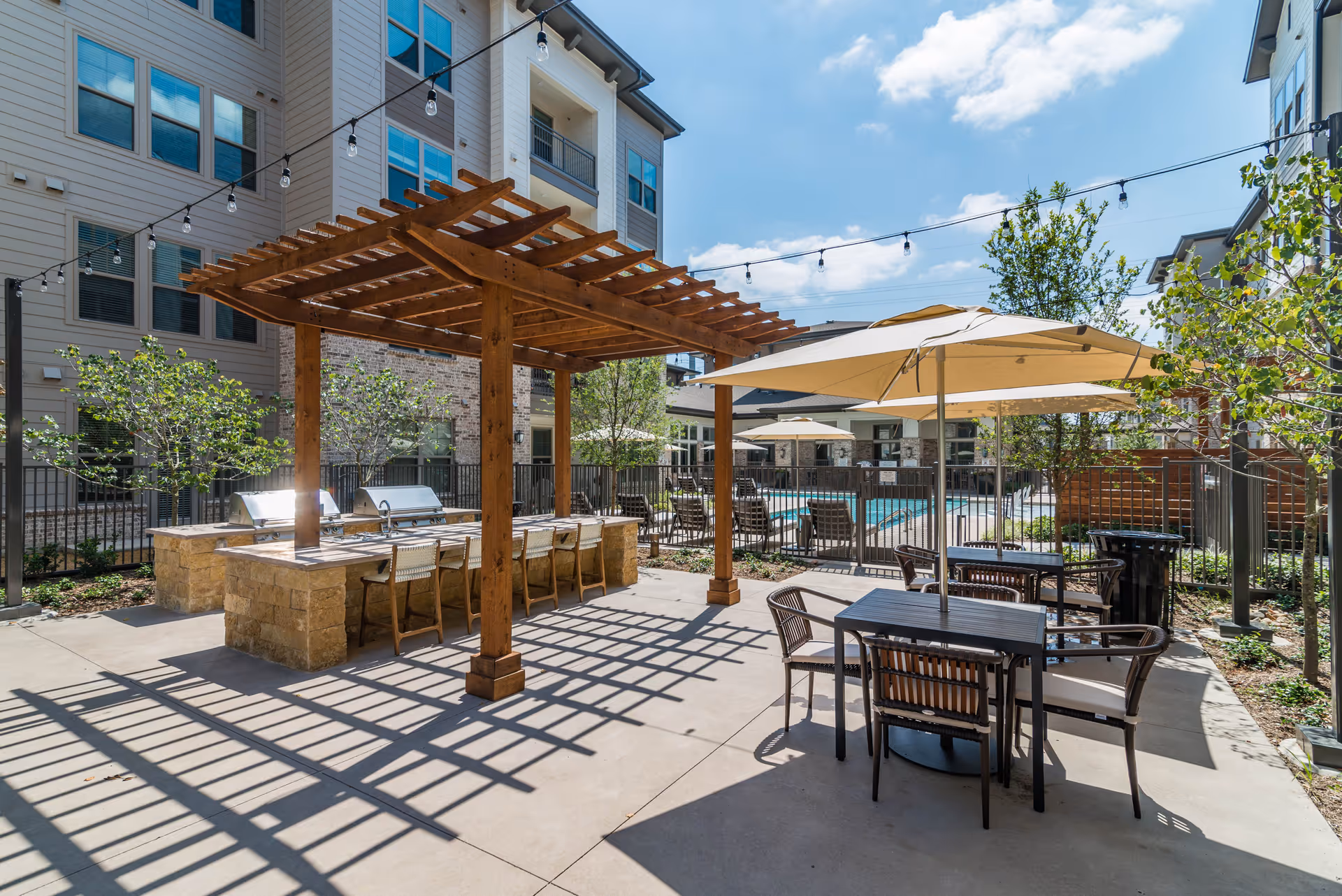 Outdoor patio area at Album Keller Ranch featuring a wooden pergola with built-in grills and bar seating, tables with umbrellas, lounge chairs near a fenced swimming pool, surrounded by multi-story residential buildings and greenery under a partly cloudy sky.