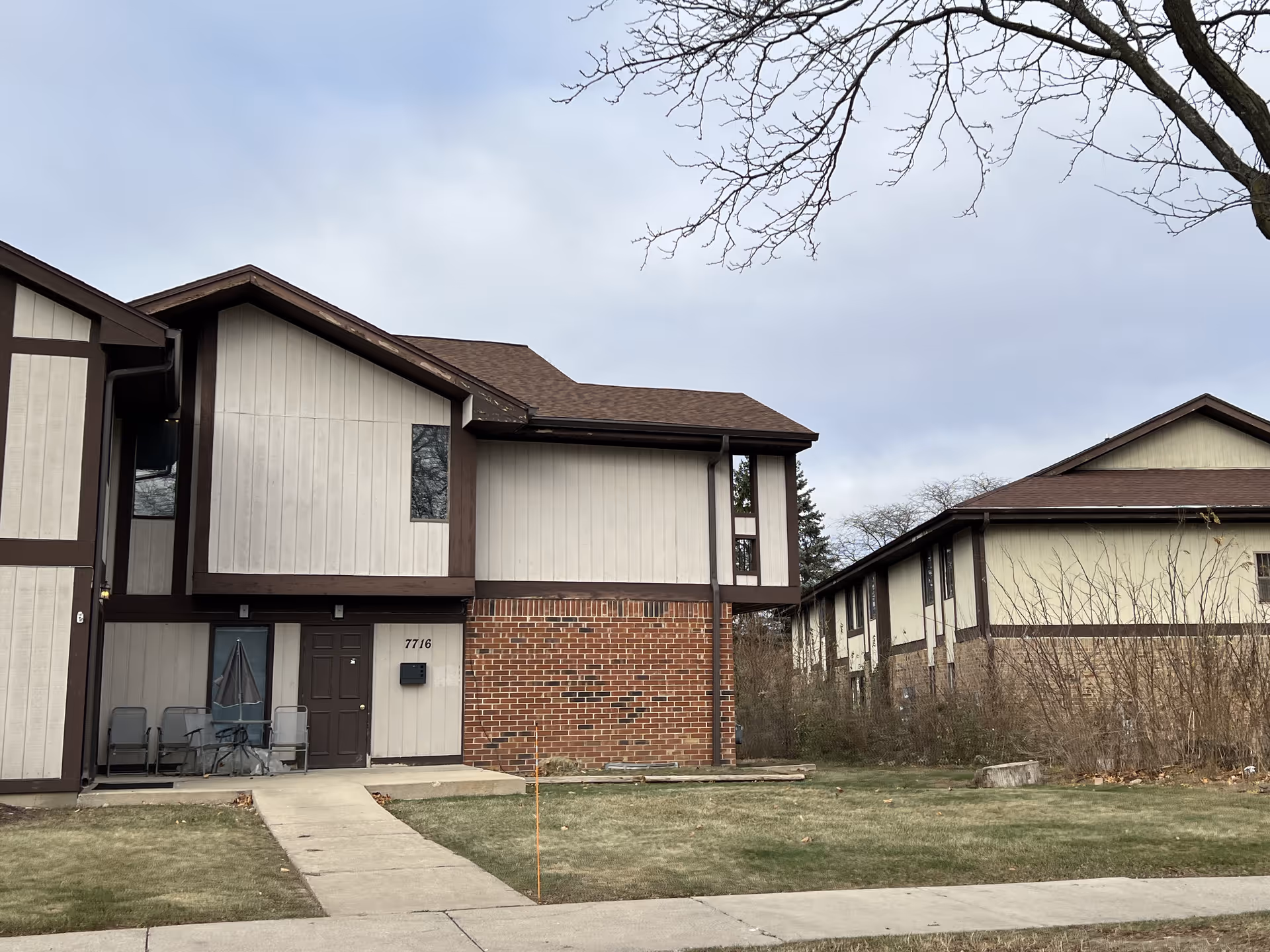 Exterior view of a two-story residential building with beige siding and brown trim. The building has a small concrete walkway leading to a front door with a covered patio area furnished with chairs and a table. There is a grassy lawn in front and leafless trees in the background under a cloudy sky.