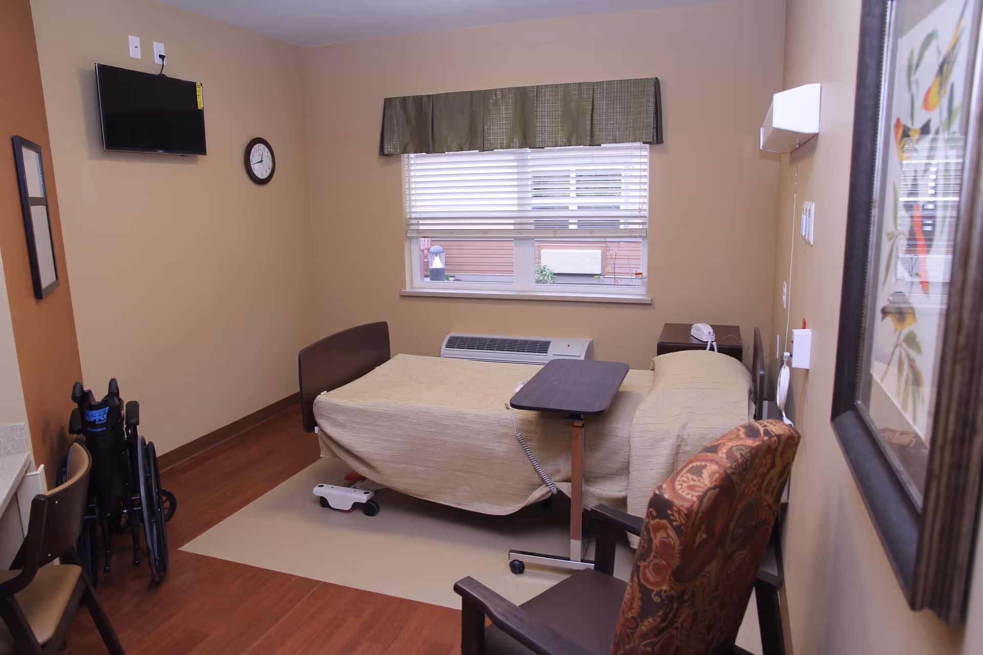 A small, neatly arranged room with a single hospital-style bed covered with a beige blanket. There is a brown patterned armchair near the bed, a small overbed table, a wheelchair folded and placed near the corner, a wall-mounted TV, a clock, and a window with blinds and a green valance. The walls are painted beige and light brown, and there is a framed picture on the wall.
