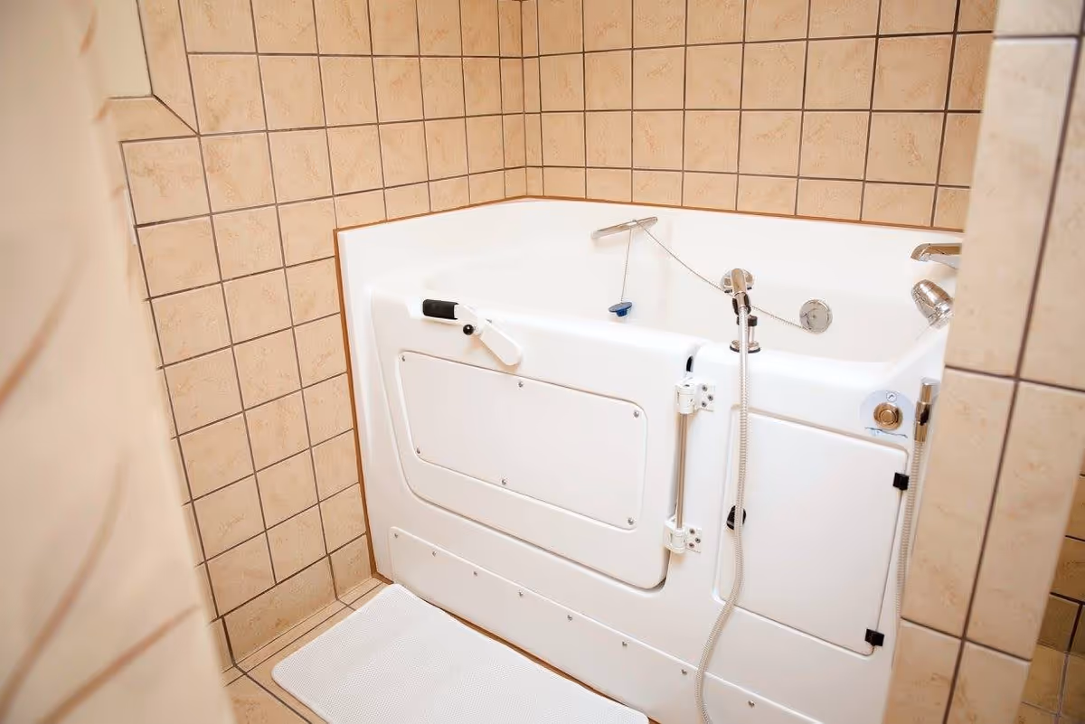 A white walk-in bathtub with a grab handle and handheld shower against beige tiled walls.