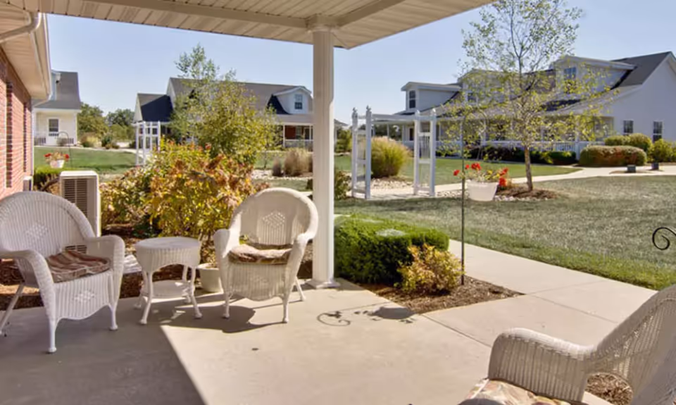 Covered patio with white wicker chairs overlooking a landscaped courtyard and nearby cottage-style senior living buildings.