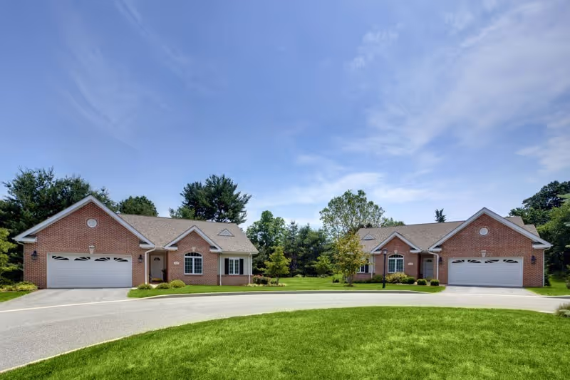 Two single-story brick homes with attached garages facing a curved driveway and landscaped lawn under a blue sky.