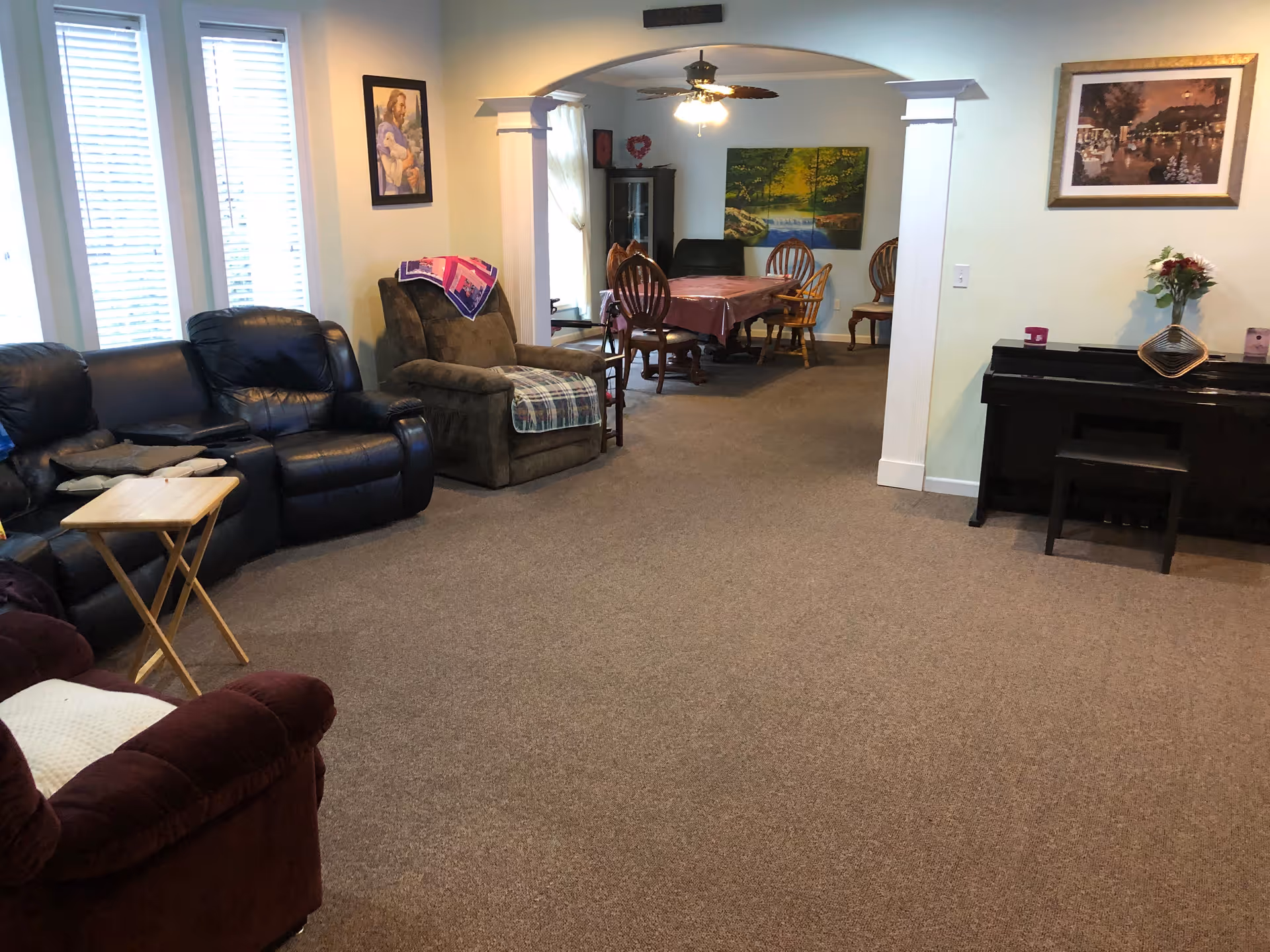 A cozy living room area with multiple armchairs and a small wooden folding table. The room has beige carpet and light-colored walls with framed artwork. In the background, there is a dining area with a table covered by a pink tablecloth and several wooden chairs. A black piano with a small bench and a flower vase is positioned against the right wall.
